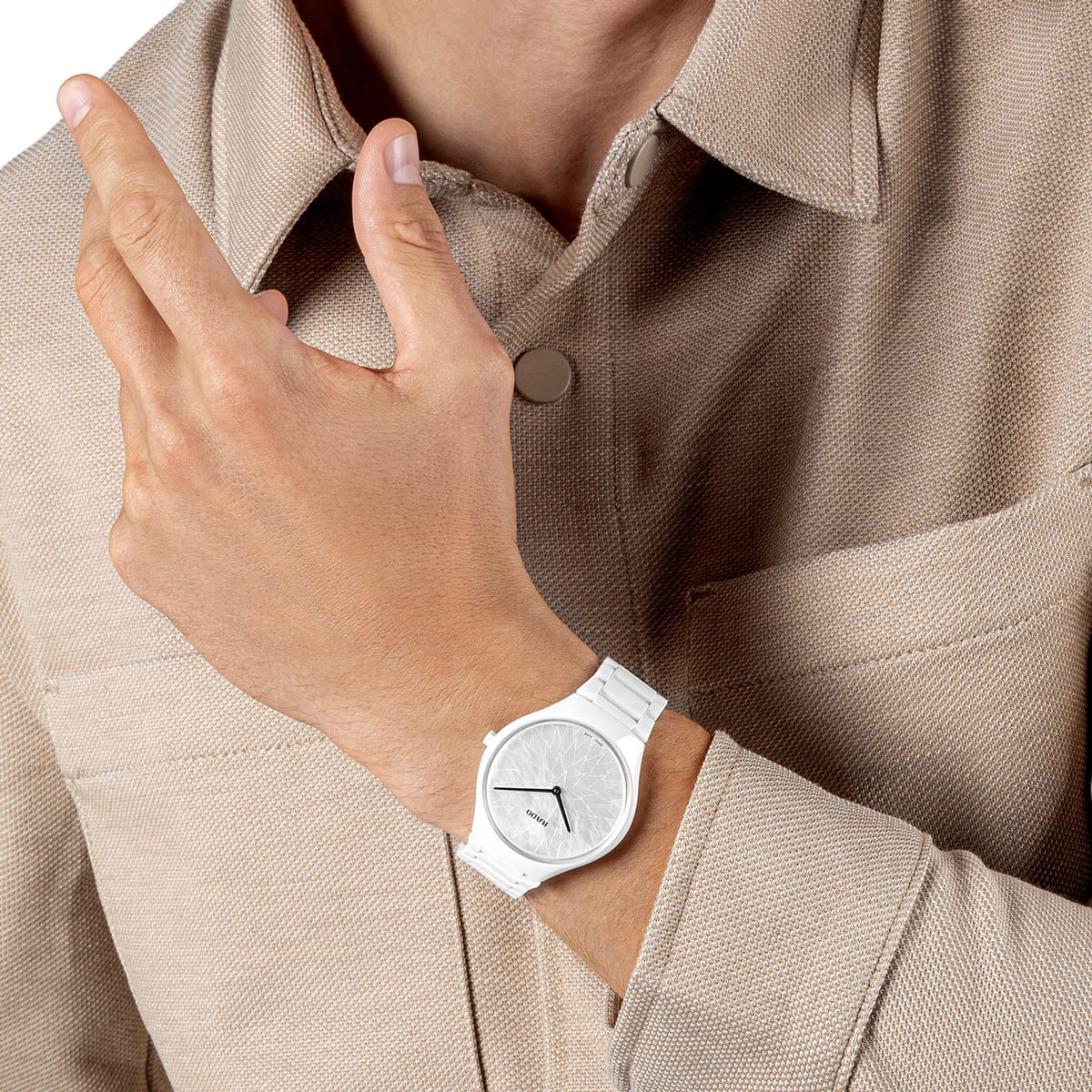 RADO © 2026 Eine Person, die eine beigefarbene strukturierte Jacke und eine minimalistische weiße Rado True Thinline Armbanduhr trägt und die Hand in der Nähe des Kragens hebt.