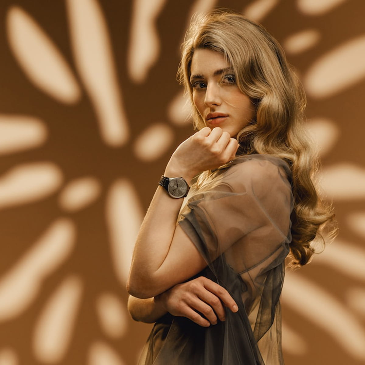 RADO © 2026 Eine Frau mit langem, gewelltem blondem Haar posiert selbstbewusst in einem transparenten, taupefarbenen Oberteil und präsentiert eine stilvolle Rado True Thinline Armbanduhr. Der Hintergrund zeigt ein warmes, abstraktes Muster mit weichen, leuchtenden Lichtformen.