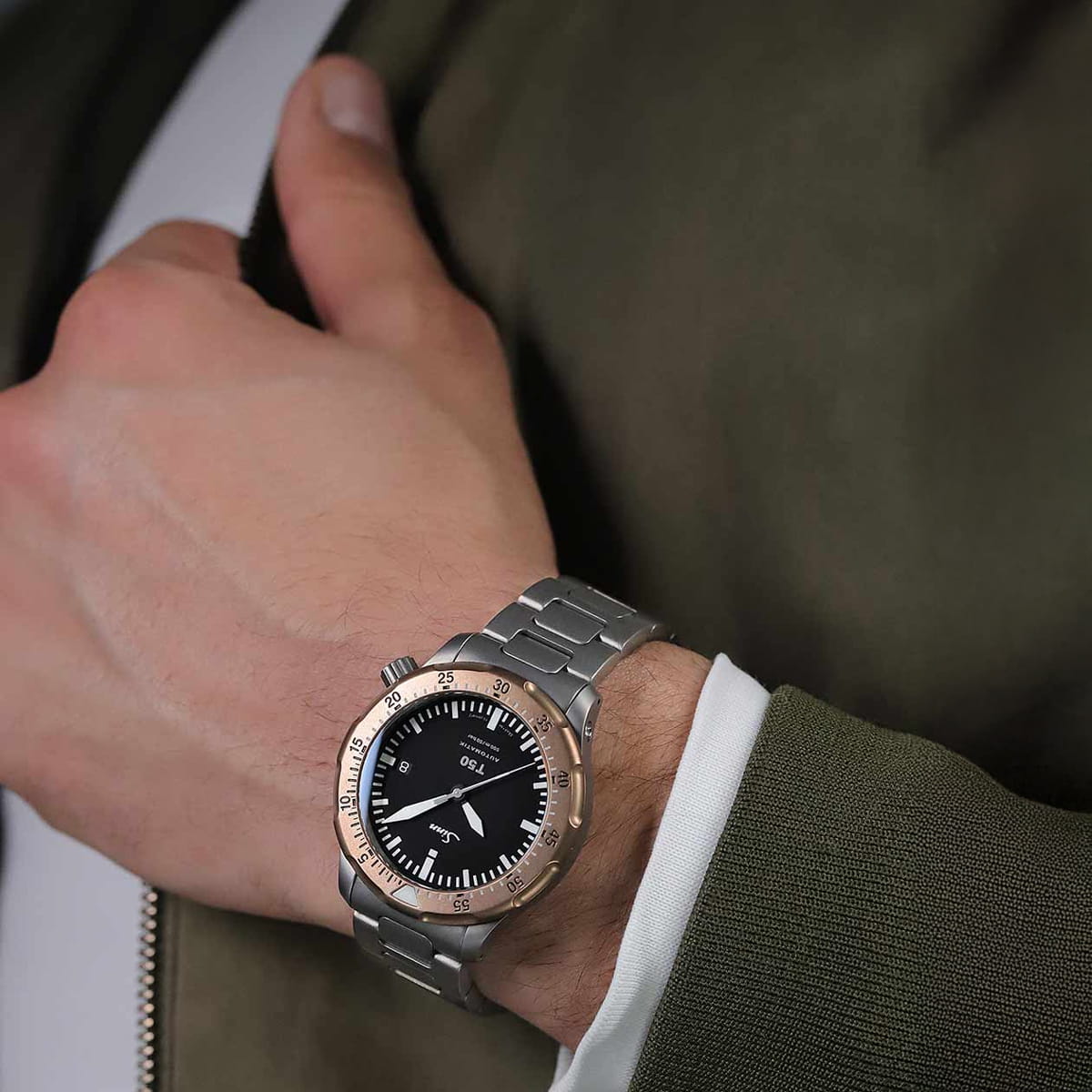 Eine Person in olivgrüner Jacke und weißem Hemd trägt eine Sinn Spezialuhren in Silber und Roségold mit schwarzem Ziffernblatt und weißen Markierungen und stützt ihre Hand auf die Brust.