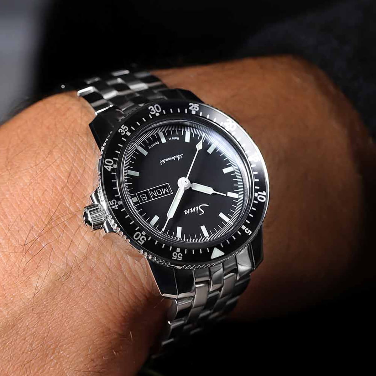 Eine Nahaufnahme des Handgelenks einer Person, die eine Sinn Spezialuhren-Armbanduhr aus Edelstahl mit schwarzem Zifferblatt, silbernen Indexen, einer drehbaren Lünette und einem Metallarmband trägt. Die Uhr zeigt den Tag und das Datum an.