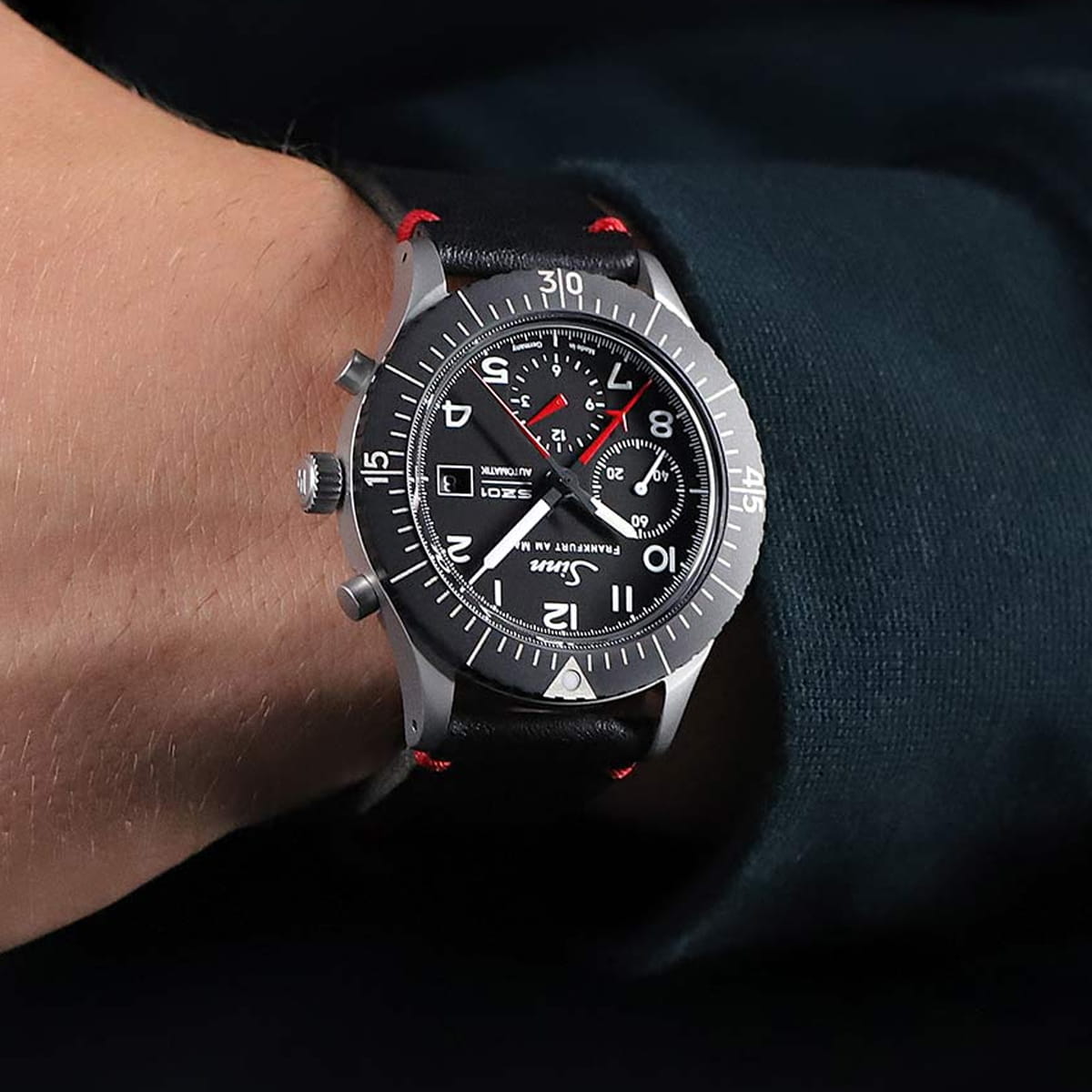 Eine schwarze Sinn Spezialuhren Armbanduhr mit rundem Zifferblatt und roten Akzenten wird am Handgelenk einer Person getragen. Die Uhr verfügt über drei Hilfszifferblätter, große weiße Ziffern und ein schwarzes Lederarmband mit roten Nähten.