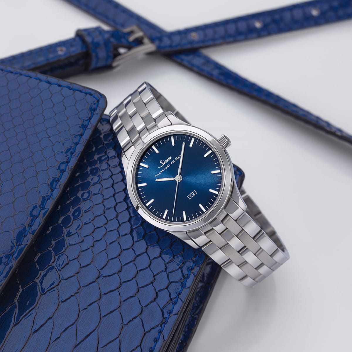 Eine silberne SINN Damenuhren-Armbanduhr mit blauem Ziffernblatt und Metallband ruht auf einer blauen, strukturierten Lederbörse, wobei im Hintergrund ein passendes blaues Armband sichtbar ist.