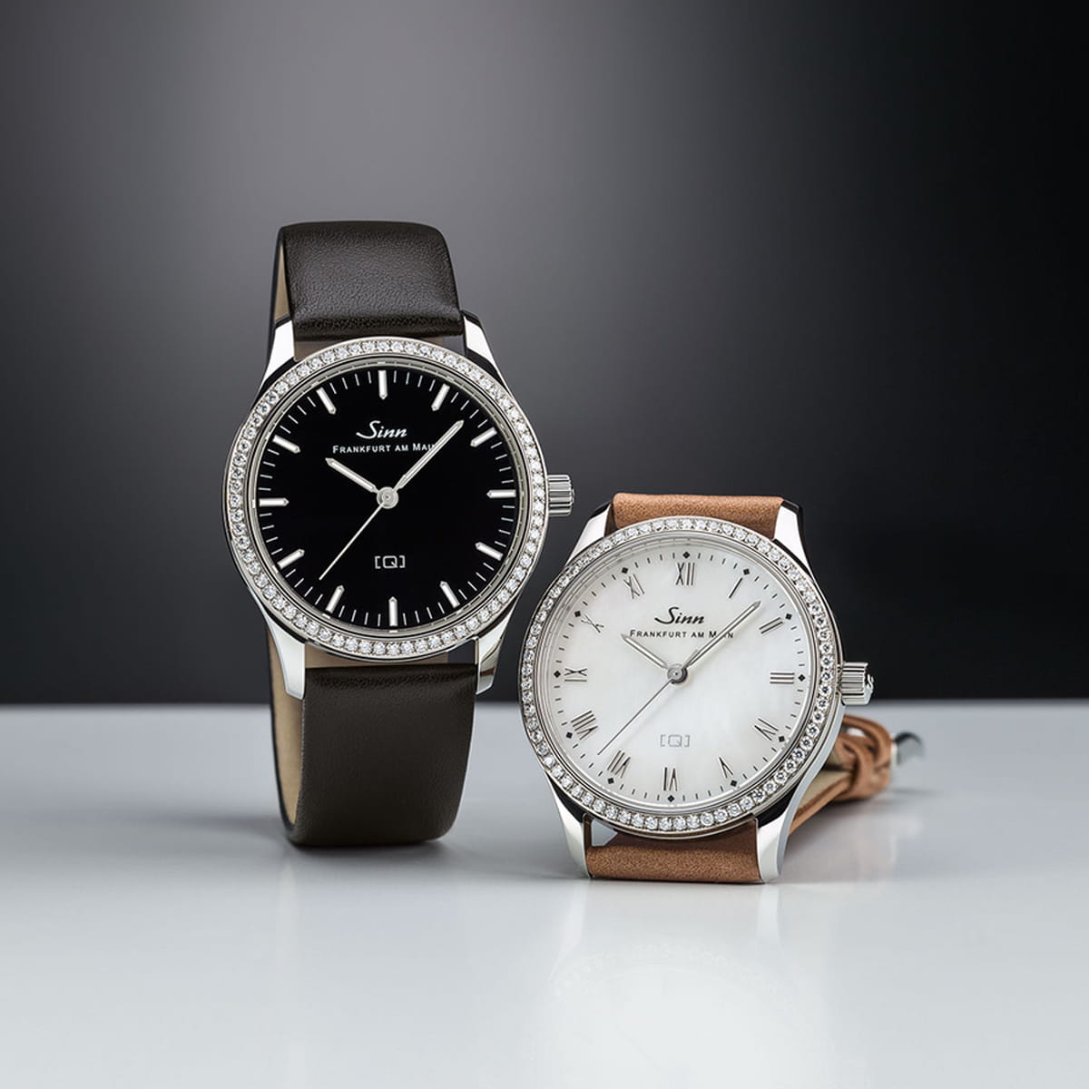 Zwei elegante Sinn Spezialuhren mit Diamantlünetten: eine mit schwarzem Zifferblatt und schwarzem Lederarmband, die andere mit weißem Zifferblatt und römischen Ziffern und hellbraunem Lederarmband, beide vor dunklem Hintergrund.