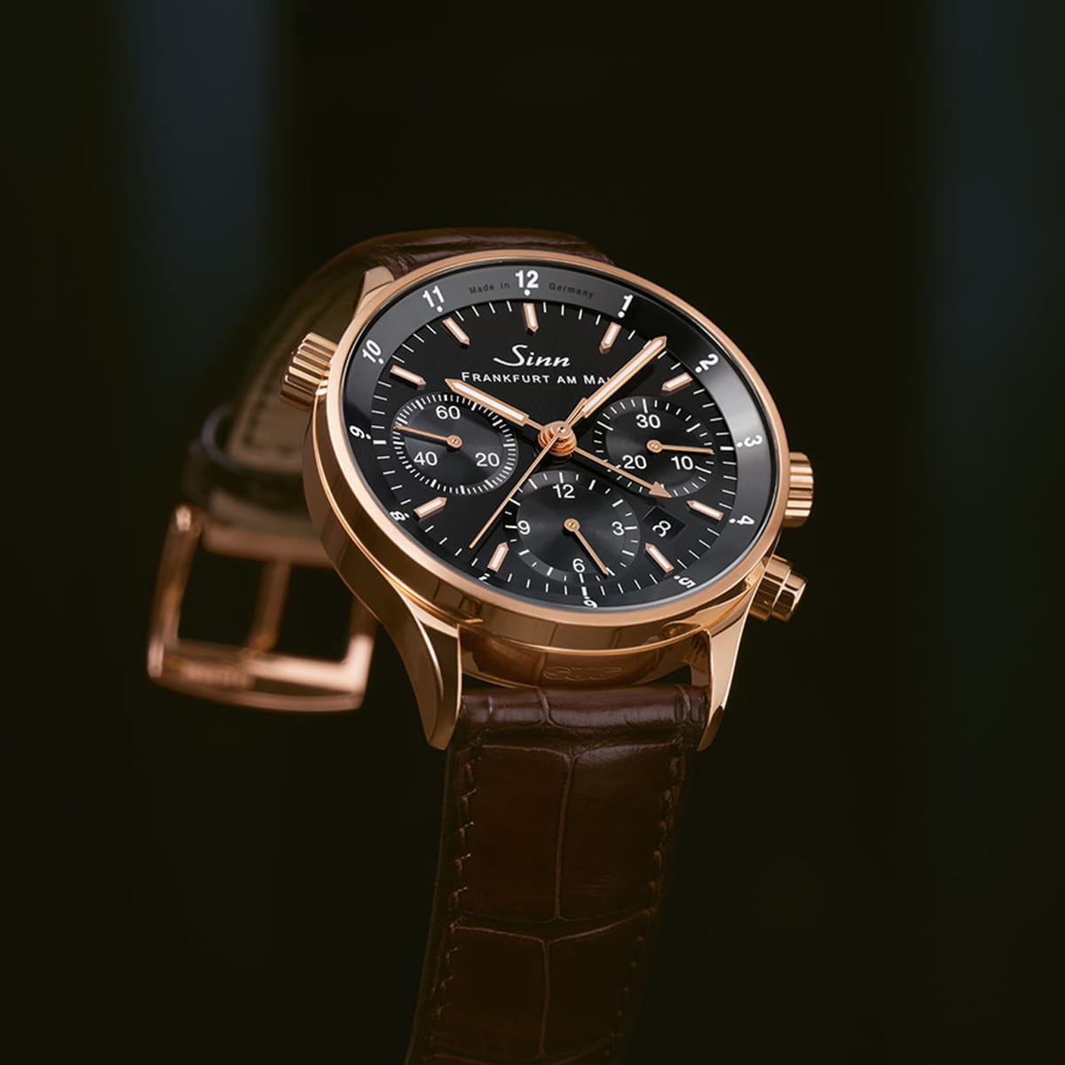 Eine goldene Sinn Frankfurter Finanzplatzuhren Armbanduhr mit schwarzem Zifferblatt, Chronographenzifferblättern und braunem Lederarmband wird vor einem dunklen Hintergrund gezeigt. Das Branding und die Zahlen sind auf dem Zifferblatt sichtbar.