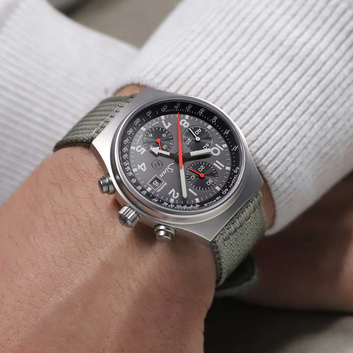 SINN Spezialuhren © 2026 Nahaufnahme einer Person, die eine Sinn-Chronographen-Armbanduhr mit grauem Armband und schwarzem Zifferblatt mit roten und weißen Details über einem weißen langärmeligen Rippenhemd trägt.