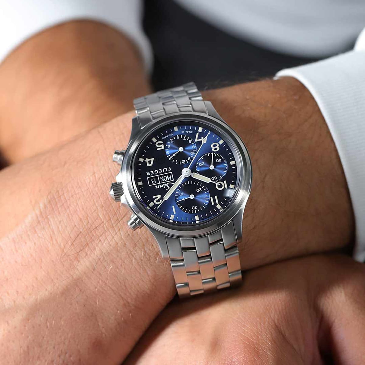 SINN Spezialuhren © 2026 Nahaufnahme des Handgelenks einer Person, die eine silberne Sinn-Chronographen-Uhr mit blauem Zifferblatt, silbernen Stundenmarkierungen und mehreren Hilfszifferblättern trägt. Die Person ist mit einem weißen, langärmeligen Hemd bekleidet.