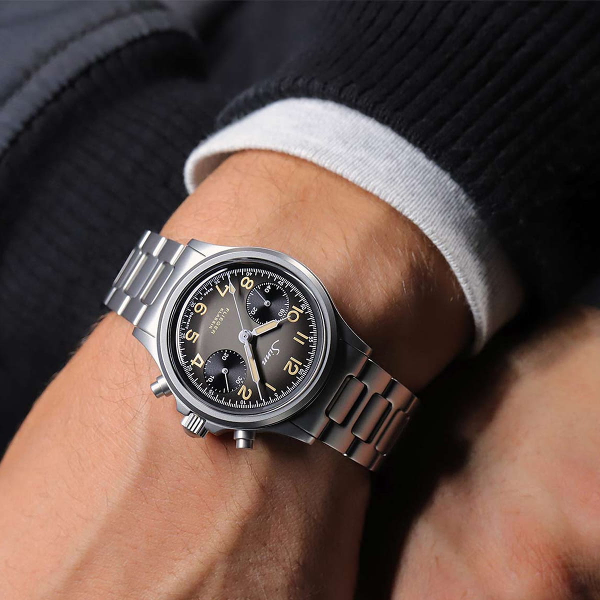 SINN Spezialuhren © 2026 Nahaufnahme des Handgelenks einer Person, die eine Sinn Chronographen-Edelstahluhr mit schwarzem Zifferblatt, zwei Hilfszifferblättern und gelben Ziffern trägt. Die Person trägt einen schwarzen Pullover über einer weißen Manschette.