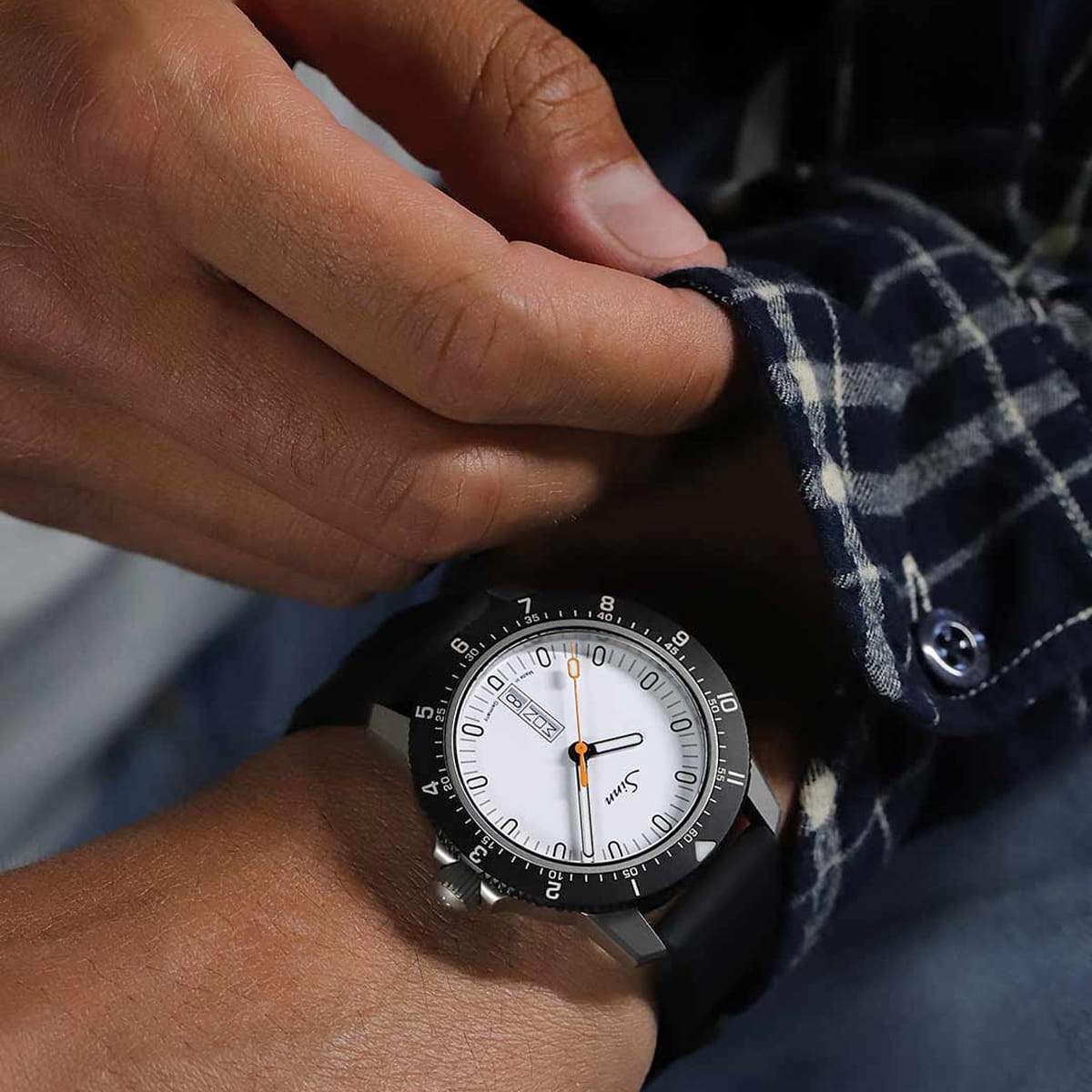 SINN Spezialuhren © 2026 Eine Person rückt die Manschette eines blau karierten Hemdes zurecht, während sie eine große, silberne Sinn Instrumentelle Uhren Armbanduhr mit weißem Zifferblatt und schwarzem Armband am linken Handgelenk trägt.