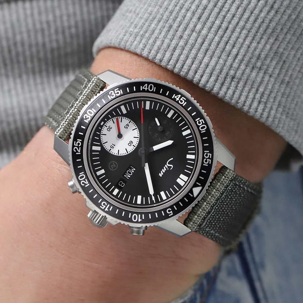 Nahaufnahme einer Person, die eine schwarze Sinn Taucheruhren-Armbanduhr mit einem grauen Stoffarmband trägt. Das Zifferblatt weist weiße Stundenmarkierungen, ein kleines weißes Hilfszifferblatt und silberne Knöpfe auf. Der Träger ist mit einem grauen Pullover und Jeans bekleidet.