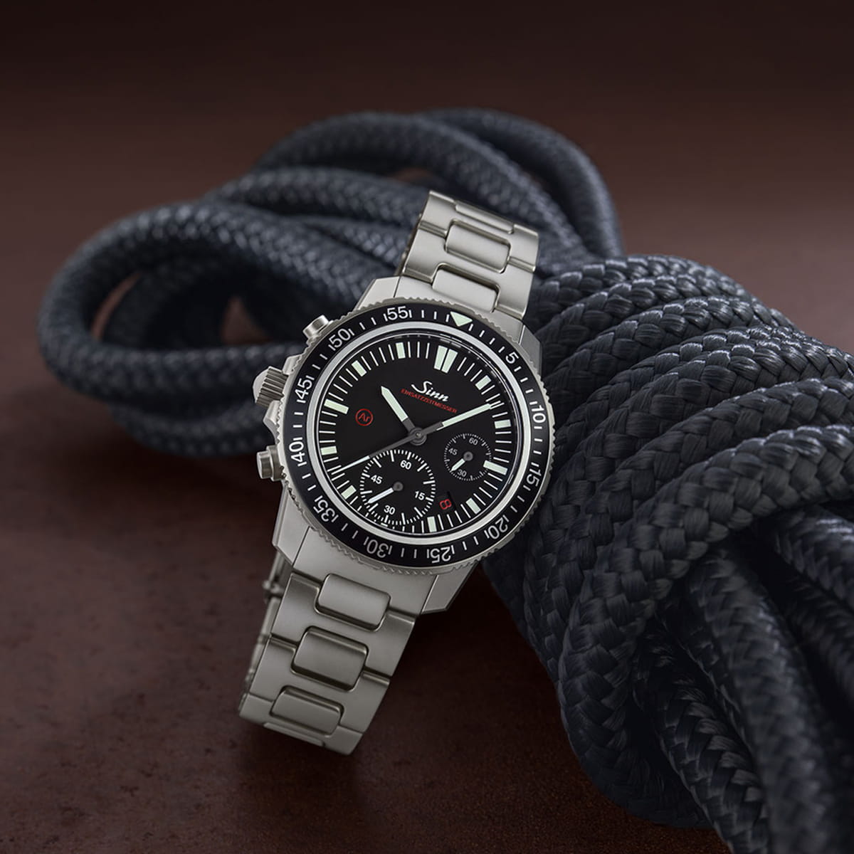 Eine Sinn Taucheruhren-Armbanduhr aus Edelstahl mit schwarzem Ziffernblatt und mehreren Hilfszifferblättern wird auf einem aufgewickelten schwarzen Seil vor einem braunen Hintergrund präsentiert.