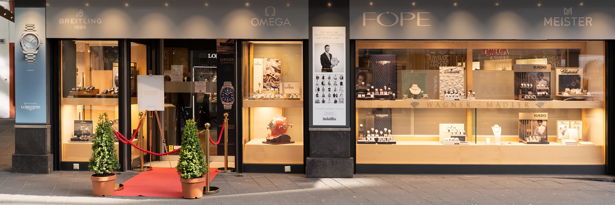 © Juwelier Wagner-Madler e.K. Ein Juwelier- und Uhrengeschäft mit eleganten Schaufenstern, rotem Teppich, goldenen Stützen und Topfpflanzen am Eingang; ein engagiertes Team präsentiert Luxusmarken wie Omega, Fope, Meister und Breitling in der Schaufensterfront.