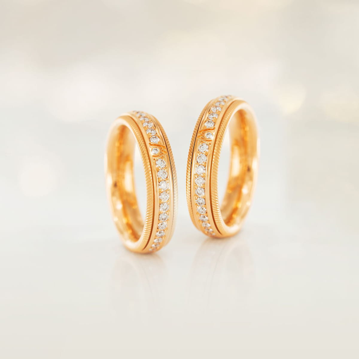 Zwei goldene Eheringe mit einer Reihe kleiner, funkelnder Diamanten in der Mitte, aufrecht stehend auf einer weichen, hellen Oberfläche mit verschwommenem Hintergrund.