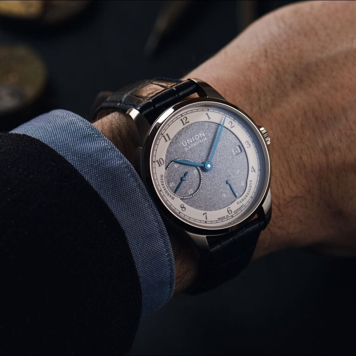 Nahaufnahme eines Handgelenks mit einer eleganten Union Glashütte 1893 Analog-Armbanduhr mit silbernem Zifferblatt, blauen Zeigern und schwarzem Lederarmband. Die Person ist mit einem dunklen Anzug und einem hellblauen Hemd bekleidet.