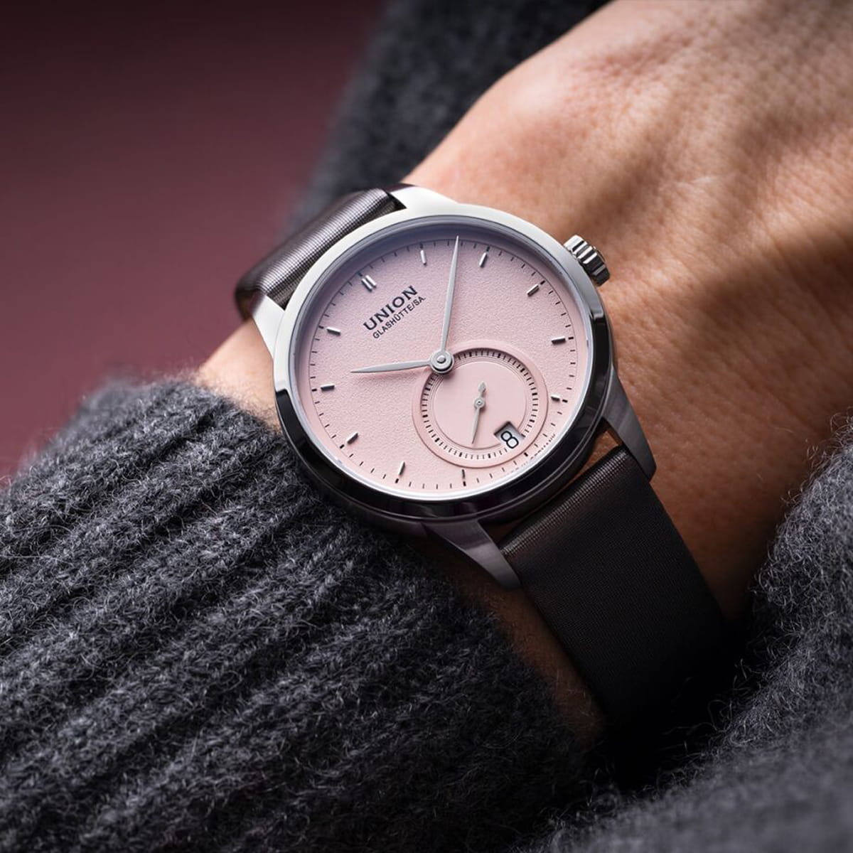 Eine Person, die eine silberne Armbanduhr Union Glashütte 1893 mit einem blassrosa Zifferblatt, schwarzen Indexen, einer kleinen Sekundenanzeige und einem schwarzen Lederarmband trägt, bekleidet mit einem dunkelgrauen, strukturierten Pullover.