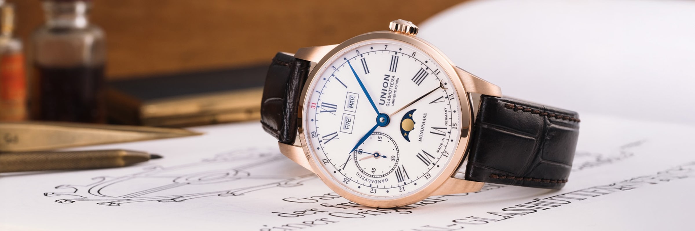 Eine luxuriöse Union Glashütte 1893 Armbanduhr mit weißem Zifferblatt, goldenem Gehäuse, schwarzem Lederarmband, römischen Ziffern und einer Mondphasenanzeige ruht auf einem aufgeschlagenen Buch mit handschriftlichem Text. Im Hintergrund sind ein goldener Füller und Tintenfässer zu sehen.