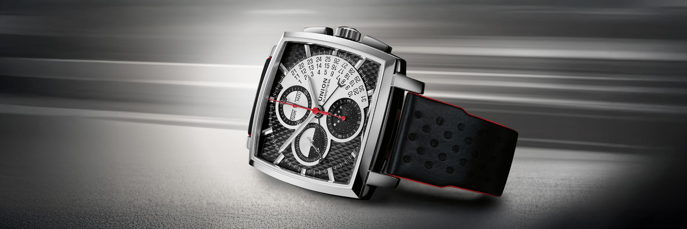 Eine luxuriöse Union Glashütte Averin Armbanduhr mit quadratischem Zifferblatt, schwarzem perforiertem Armband, silbernem Gehäuse und detailliertem schwarz-weißem Zifferblatt mit roten Akzenten vor einem verschwommenen grauen Hintergrund.