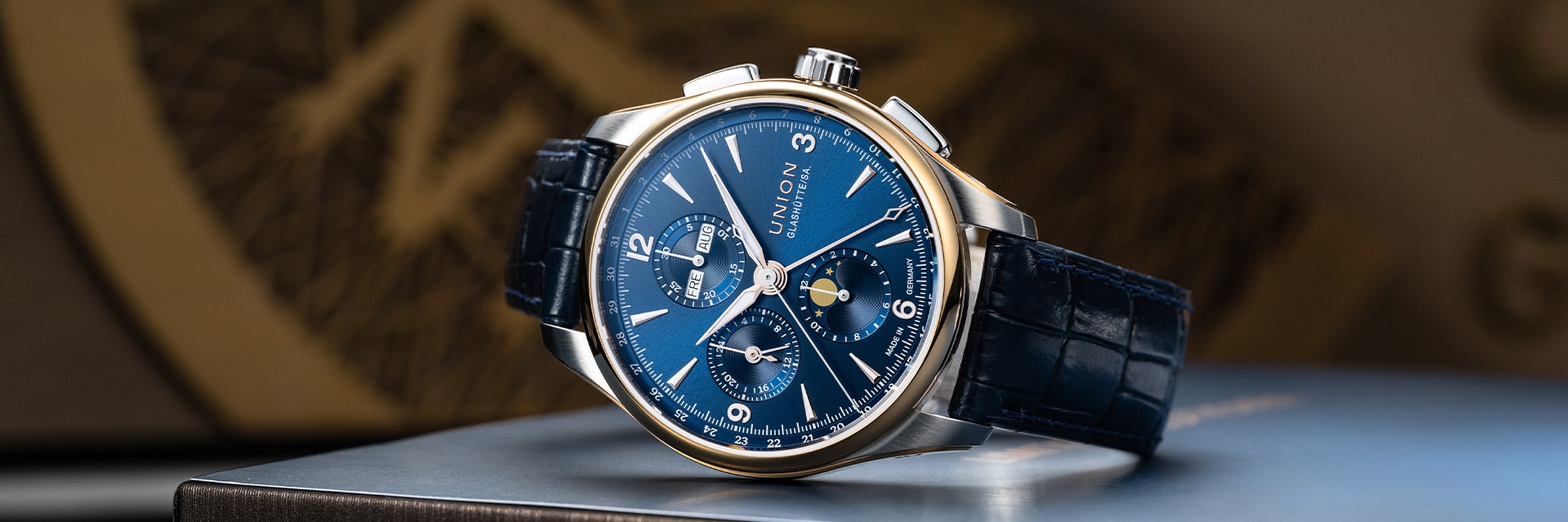 Eine luxuriöse Union Glashütte Belisar Armbanduhr mit blauem Lederarmband und blauem Zifferblatt mit mehreren Hilfszifferblättern, silbernen Indexen und einer goldfarbenen Lünette, die auf einer Metalloberfläche angebracht ist.