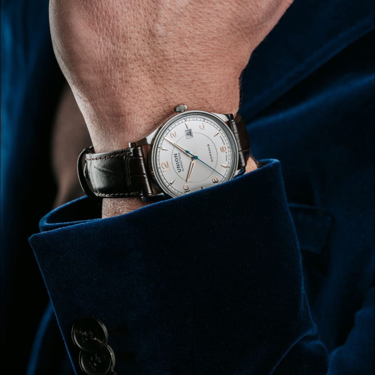 Eine Person, die eine dunkelblaue Samtjacke trägt, zeigt eine silberne Union Glashütte Noramis Armbanduhr mit einem braunen Lederarmband und einem weißen Zifferblatt mit schwarzen Ziffern und blauen Zeigern.