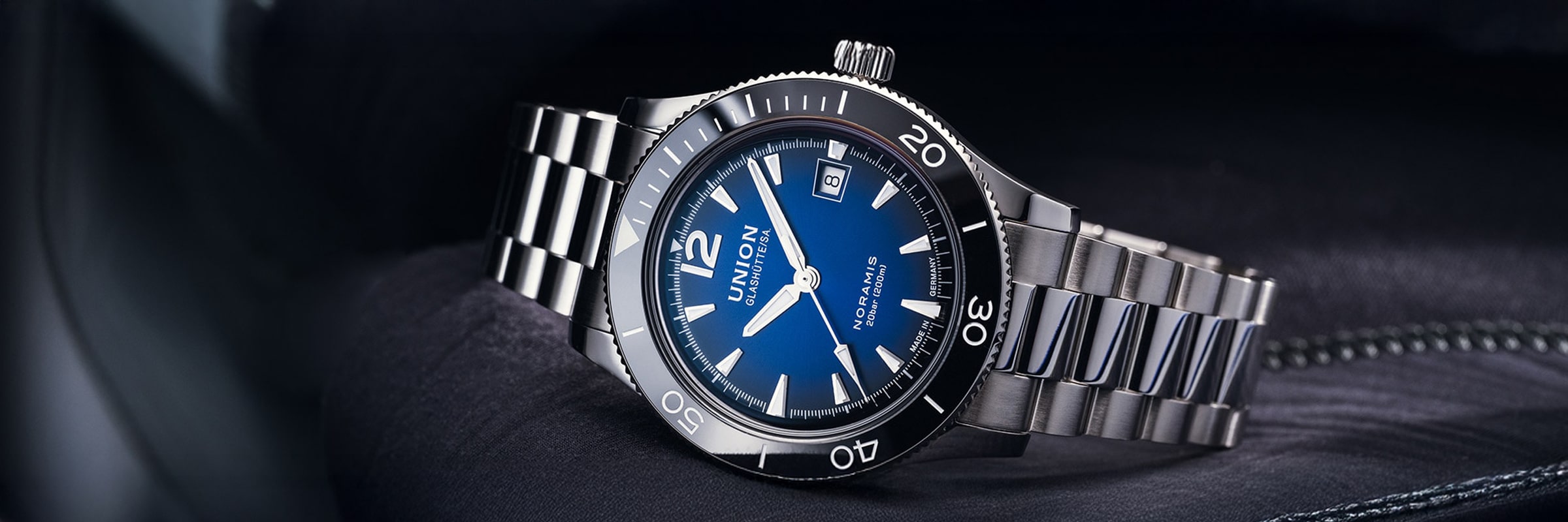 Nahaufnahme einer Union Glashütte Noramis Luxusarmbanduhr mit blauem Zifferblatt, silbernen Indexen, drehbarer Lünette und silbernem Metallarmband, die auf einer dunklen Stoffoberfläche liegt. Die Uhr zeigt die Zeit und das Datum an.