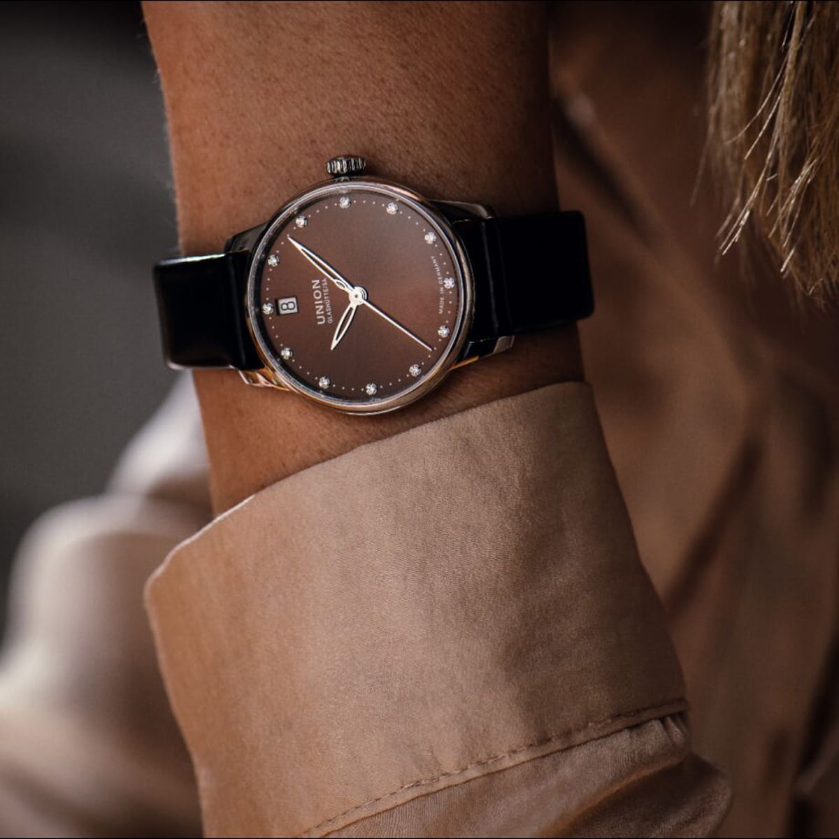 Nahaufnahme eines Handgelenks mit einer Union Glashütte Seris-Uhr mit braunem Zifferblatt, Diamantindexen, Datumsfenster und schwarzem Lederarmband. Die Person ist mit einem beigen Hemd bekleidet.