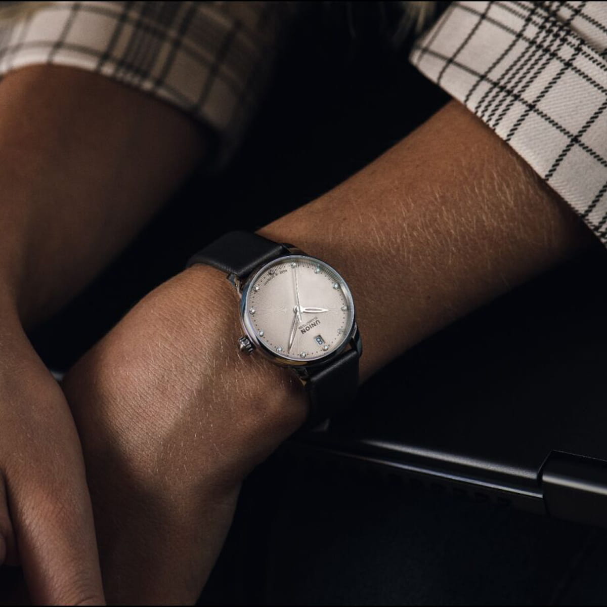 Nahaufnahme einer Person, die eine silberne Union Glashütte Seris-Armbanduhr mit schwarzem Armband und weißem Zifferblatt trägt und ihr Handgelenk auf einer dunklen Fläche abstützt. Die Person ist mit einem karierten Hemd bekleidet.