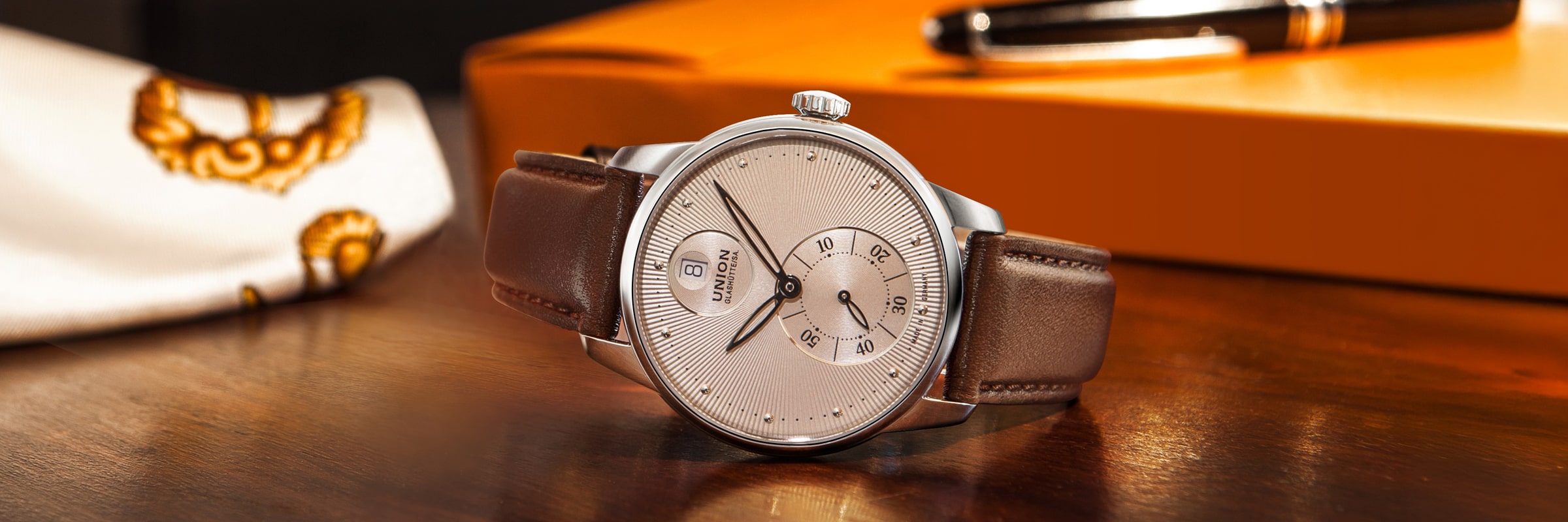 Eine silberne Armbanduhr von Union Glashütte Seris mit braunem Lederarmband ruht auf einer polierten Holzfläche. Dahinter sind ein weiß gemustertes Tuch, eine orangefarbene Schachtel und ein schwarz-goldener Stift sanft beleuchtet.