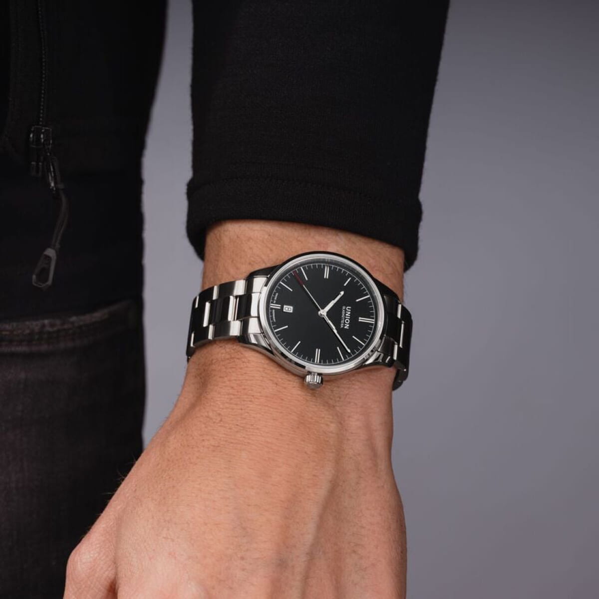 UNION GLASHÜTTE © 2026 Eine Person trägt eine Union Glashütte Viro-Armbanduhr mit schwarzem Zifferblatt, silbernen Stundenmarkierungen und Datumsanzeige. Sie ist schwarz gekleidet und steht vor einem neutralen grauen Hintergrund.