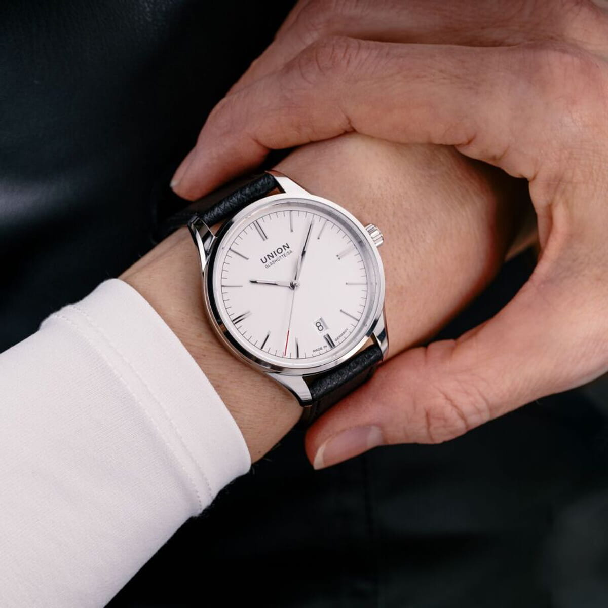 Nahaufnahme einer Person, die eine Armbanduhr mit weißem Zifferblatt, einfachen schwarzen Indexen und einem schwarzen Lederarmband einstellt. Die Uhr trägt die Marke "Union Glashütte" und hat eine Datumsanzeige.