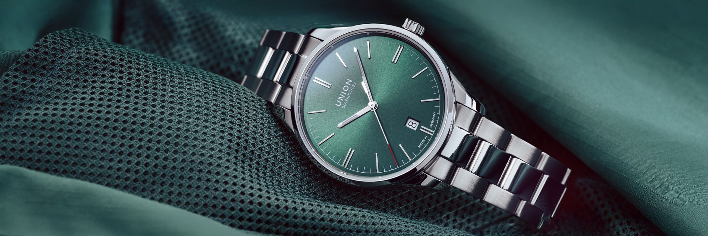 UNION GLASHÜTTE © 2026 Eine silberne Union Glashütte Viro Armbanduhr mit grünem Zifferblatt und silbernen Indexen ruht auf grünem Stoff, zeigt 10:10 Uhr an und verfügt über ein Datumsfenster bei 3 Uhr.