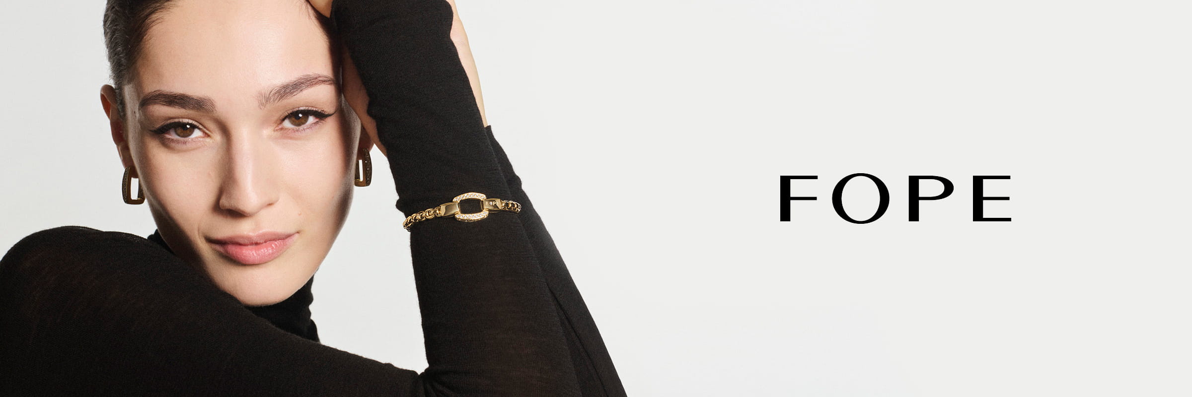 Eine Frau mit braunem Haar, die einen schwarzen Rollkragenpullover, goldene Ohrringe und ein goldenes Fope-Armband trägt, posiert mit erhobenem Arm. Neben ihr erscheint das Wort "FOPE" in schwarzer Schrift auf weißem Hintergrund.